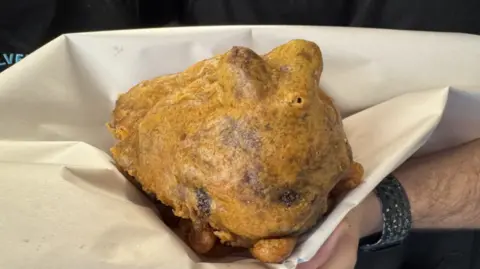 A deep fried object in golden batter held in a large sheet of white paper.