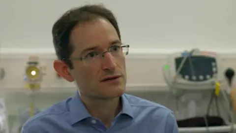 A man with glasses wearing a light blue shirt. In the background to the right a medical machine can be seen