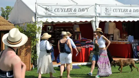 Spectators take a break from the action to do a spot of shopping. Ladies wearing summer hats and dresses eye up some clothes on a stand by London based Cuyler and Davy.