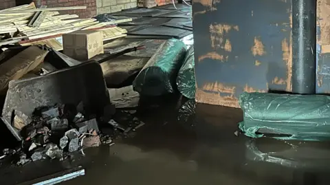 The floor of a mill is covered in water with wood and boxes toppled up in the far left corner. 