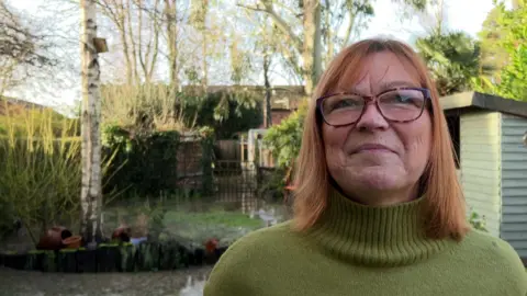 Susannah Gill giving a BBC interview in her sewage-flooded garden. It is a sunny day.