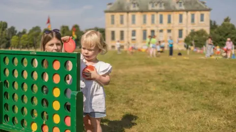 National Trust A woman with light brown hair and a toddler with blonde hair and a while top and denim shorts play a game Connect Four on a large version of it, complete with large red and yellow tokens. The child is holding two red tokens and is about to slot one into the green board. They are on the grass with a large stately home in the background and other blurred people playing other games.