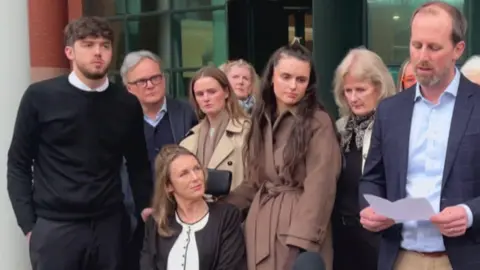 BBC Blonde haired Trudi Burgess, looks up at her brother Charlie from her wheelchair as he reads out a statement to the press outside court. She is surrounded by friends and family