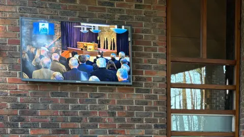 PA Media A TV screen fixed to the outside wall of the crematorium shows four rows o people, with the coffin on a plinth and a photo of Tony Martin on a TV screen at the top of the altar.