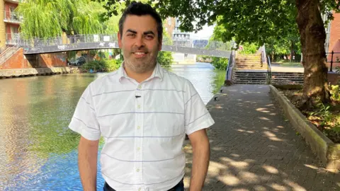 A smiing Eugene Mohareb standing under the shade of a tree on a footpath alongside the Kennet and Avon canal as it passes through Reading town centre. There's a pedestrian footbridge across the canal just behind him.