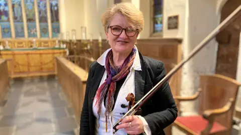 Marion Fleetwood wearing glasses, standing up . Behind her are rows of wooden pews with lamps on them