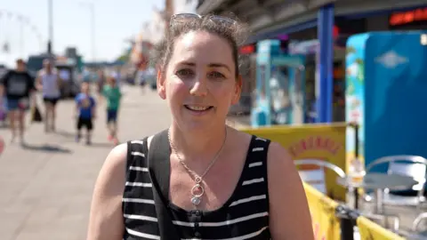 Qays Najm/BBC Louise Jones looks directly at the camera. She is standing on a seaside promenade. She is smiling and wearing a black vest top with white horizontal stripes and a necklace. She has her hair tied back and sunglasses perched on top of her head.