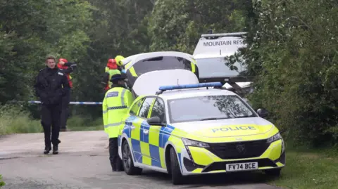 PA Media A police car and officers with a van behind in a lane with trees.