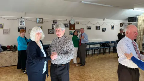 George Carden/BBC Club members dancing in pairs in the building's hall. There are pictures hung up on the back wall of the club's history and the floor is a wooden ball room type floor.