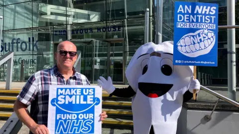 Matt Marvel/BBC Mark Jones smiles the camera while standing outside Suffolk County Council's headquarters. He is bald and wears sunglasses along with a chequered shirt. He stands next to another person dressed in a tooth outfit. Both are holding up placards calling for more NHS dentists