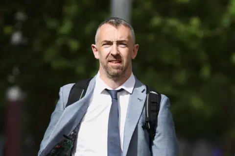 PA Media Daithí McKay has salt and pepper coloured hair and a beard and is wearing a light grey suit, white shirt and dark coloured tie   