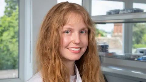 Dr Lindell is in a lab with windows behind her. She is smiling at the camera while wearing a white lab coat. She has long ginger hair and light blue/ green eyes. 