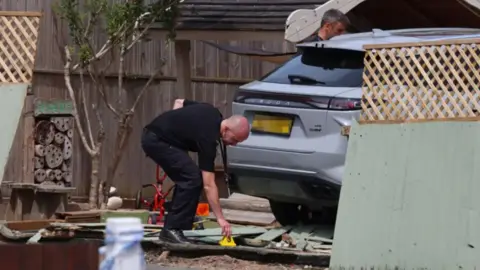 Reach plc A man leans down to place a yellow marker of evidence on the ground amid rubble and fencing after a silver car beside him crashed through a fence leading to a nursery playground.