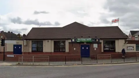 A single storey building, with a navy blue double door and three large windows. There is a banner saying "Welcome back" above the door, and an England flag flying from a flag pole on the roof.