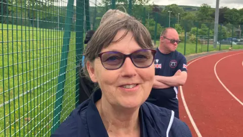 Grace Wood/BBC A woman with tied back brown hair wearing dark glasses smiles at the camera. She is standing on an athletics track