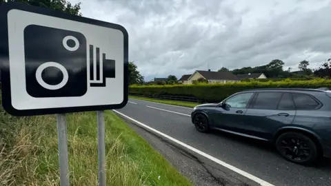 A black and white speed camera road sign on a grass verge at the side of a road, with a residential area in the background. A dark grey vehicle is travelling past the sign. 