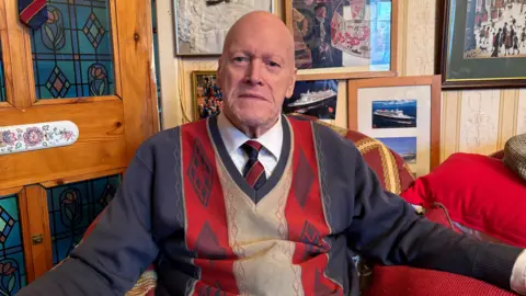 Michael is wearing a blue and red jumper, a white shirt and a blue and red tie. He is sitting in an armchair in a living room. There are pictures in frames on the wall behind him.