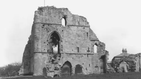 Getty Images The image is a black-and-white photograph of castle ruins on grass, with open sky behind it. The main building is a large, rectangular stone structure. The front façade features large, arched openings. Above these there is a tall, pointed Gothic-style arch window partially filled with debris and vegetation.