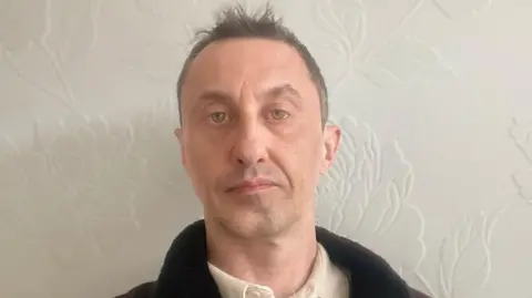 David Robinson is photographed indoors against a pale wall with a subtle floral pattern. He is shown from the shoulders up, wearing a light shirt and a dark jacket. The lighting is soft and even, and the background is uncluttered.