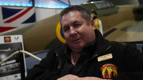 A man stands in front of the Hurricane and pilot mannequin. He has short black, greying hair and is wearing a black polo shirt and a black collared fleece with a gold name badge and a museum logo. A black and white photo and a Union flag can be seen in the background.