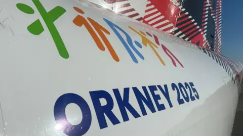 Livery on the side of an airplane which reads Orkney 2025 and has five stickmen in different colours taking different poses. There is also a tartan pattern on the tail.