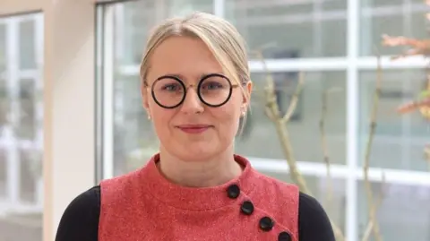 A woman with blonde hair is wearing round, black glasses and a pink top with black sleeves and black buttons on one shoulder. She is standing indoors near a set of large windows.