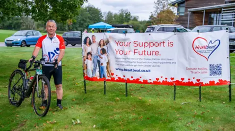 Heartbeat Nigel Brown standing on a patch of grass holding his bicycle. He is wearing a t-shirt with the logo for the charity Heartbeat. He stands next to a white banner with the charity's logo on it. The banner says "Your support is your future" and has a picture of a family on the left hand side.