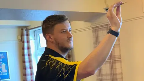 Samuel Caswell A man wearing a black and yellow shirt holds his arm up, holding a dart between his finger and thumb. There are plaid curtains behind him and a window.