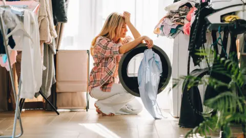 Woman with hand on her head crouches in front of a washing machine with clothes hanging up around her.