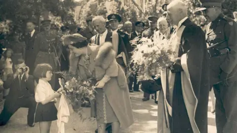 Mark Lamerton collection Several people, some in military uniform, watch as the Queen Mother receives flowers from a little girl.  The image is black and white.  