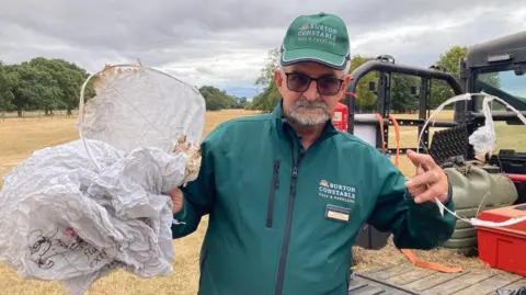 Wildlife officer Dave Nassau. He is wearing a green jacket and cap and is holding the remains of two sky lanterns. There is an expanse of brown-coloured grassland behind him.