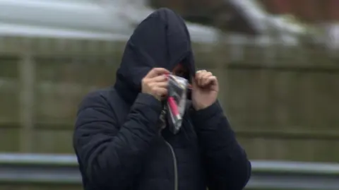 A woman is walking past the camera. she is wearing a black coat with the hood up almost entirely obscuring her face. She is holding a clear plastic bag in her hand with a pink coloured object inside.