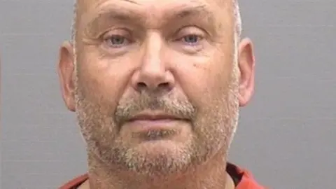 A police custody photo of a bald man with grey stubble and wearing an orange sweatshirt. The photo is taken against a plain grey backdrop and he is looking, unsmiling, at the camera.