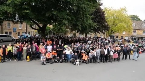 Mark Merrett A crowd of motorbike riders gather in a town square, in front of trees and Cotswold stone buildings