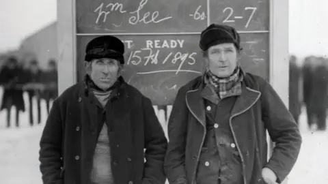 Cambridgeshire Archive A black and white photo of two middle-aged men wearing hats and layers of dark clothing, standing against a blackboard with chalked results on it. In the background, the ground is white and dark shadowy figures stand.