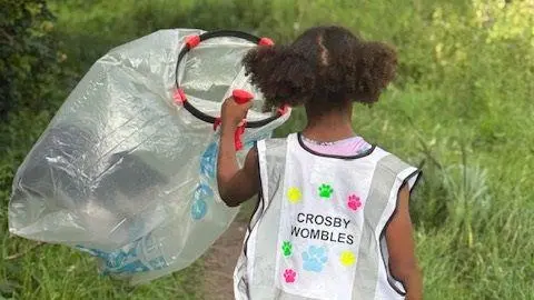 Crosby Wombles A young girl is walking in a field with a large plastic bag. She is wearing a safety vest which has the words Crosby Wombles on the back.
