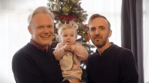 Two men hold their baby daughter in front of a Christmas tree