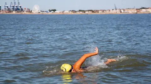 Jon Wills A woman wearing a yellow swimming cap swims in the North Sea, setting off from Felixstowe in Suffolk, which can be seen in the background on a sunny day