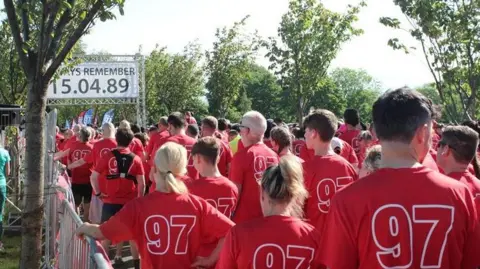 Phillip France A crowd of runners in 97 shirts wait at the start with the sign saying Always Remember 15.04.89 above. Trees are around them in the park.