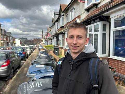 A man, who is wearing a black jacket and carrying a rucksack, is standing in front of dozens of bins in a street lined with terraced houses and cars.