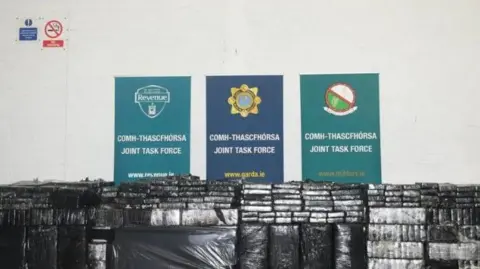 AN GARDA SÍOCHÁNA Piles of packages wrapped in black plastic in front of three signs saying 'joint task force'