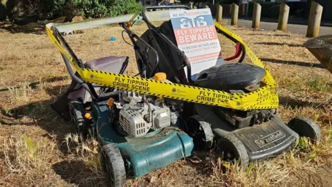 This photograph shows two discarded lawnmowers that have been positioned on a dry grassy area and surrounded with yellow crime scene tape. The mower on the left is dark green with a silver petrol engine mounted on top. It has four wheels and a black handle extending upwards.
The mower on the right is black and appears to be a Mountfield-branded mower, with the "Mountfield" logo visible on the front housing. It also has four wheels and an upright handle.
The handles of both lawnmowers are connected and wrapped together by the yellow crime-scene-style tape, creating a cordoned-off display.