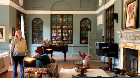 A lady stands in a beautiful sitting room with large bookcases finished with ornate glass doors 