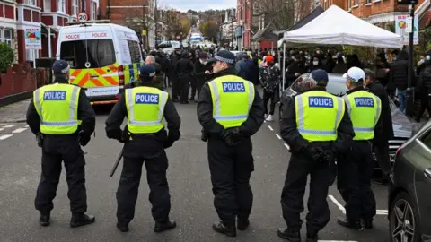 Reuters Five police officers stand in a line in a street with a police van in front of them as a crowd of people, some wearing COVID face masks, gather in front of them.