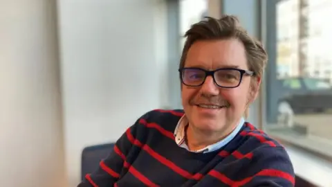 Nik Johnson wearing a navy and red striped top. He has black rimmed glasses and a moustache. 