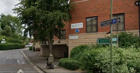 A red-brick Three Rivers District Council building stands beside a tree-lined road. Signs attached to a lamppost direct visitors to Watersmeet Theatre and the council offices. A person on a mobility scooter travels along the pavement in the foreground, with hedges and mature trees framing the scene.