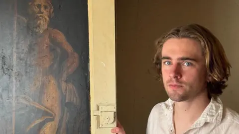 A man with brown wavy hair and a serious expression is wearing a off-white creased shirt. He is standing next to a sepia-coloured painting of an old man in a thick off-white cupboard door frame.