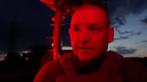 MOOSE CAMPBELL/BBC A close up picture of 'Jaeger', whose face is bathed in red light as darkness falls. He has almost no hair and wears body armour. 