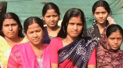 Abhishek Dey Six women residents of Rangpuri Pahari slum in India's capital city Delhi - all of them migrants from West Bengal state - wearing kurtas and sarees, stand in an open area in the neighbourhood. Behind them there's a green cloth that covers the tin wall of a tent