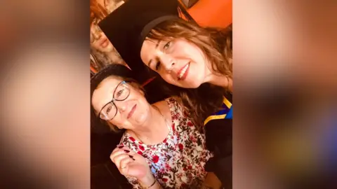 Carla Linford A selfie woman wearing a graduation cap and a gown with another woman wearing glasses and a floral dress